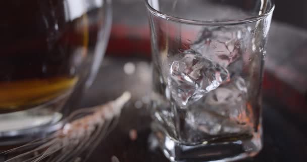 威士忌倒入一个装有冰块的杯子 站在一个老式威士忌桶上 旁边站着一个酒壶 背景是深褐色的 — 图库视频影像