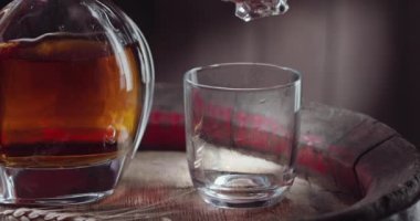 Bir buz küpü yavaşça viski bardağının üstüne dökülüyor. Eski bir viski fıçısının üstüne. İçkili bir sürahi yanında duruyor, koyu kahverengi bir arka plan. Blackmagic Ursa Pro G2. 