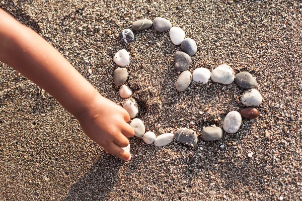 Рука малыша делает сердце из морской черепицы на песке . — стоковое фото