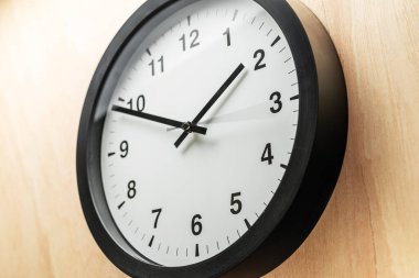 Analog duvar saati, siyah çerçeveli ve beyaz kadranlı, açık tahta duvarda asılı, saati 1: 50 olarak gösteriyor. Dakikliği, rutini ve modern iç dekorasyonu simgeleyen minimalist ve klasik bir saat..