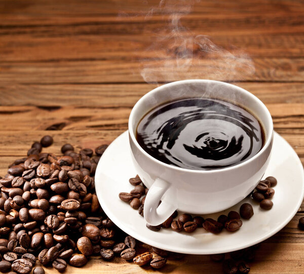 Cup of hot black coffee with steam coming out on wooden table with roasted coffee beans.