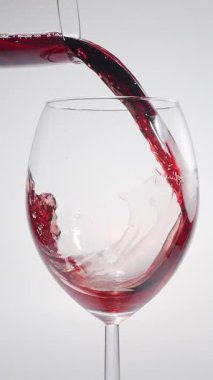 Kırmızı şarabın beyaz arka planda temiz bir bardağa dökülüşünün yavaş çekim görüntüsü. Zarif sıvı hareket, zengin renk ve şarabın dokusunun makro detaylarla yakalanması..