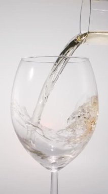 Beyaz arka planda beyaz bir bardağa dökülen beyaz şarabın yavaş çekimde yakın çekimi. Zarif sıvı hareket, zengin renk ve şarabın dokusunun makro detaylarla yakalanması..