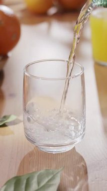 Köpüklü su plastik bir şişeden bardağa dökülüyor. Ahşap masada cam, arka planda bulanık portakallar ve elmalar..