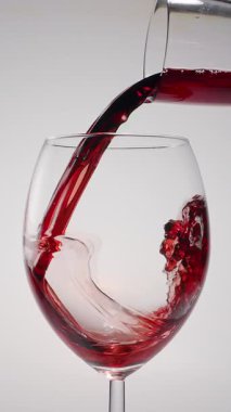 Kırmızı şarabın beyaz arka planda temiz bir bardağa dökülüşünün yavaş çekim görüntüsü. Zarif sıvı hareket, zengin renk ve şarabın dokusunun makro detaylarla yakalanması..
