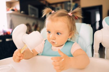 Mutfakta, masada, çocuk sandalyesinde yoğurt yiyen bir kız çocuğu.