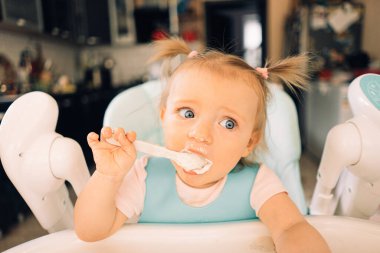 Mutfakta, masada, çocuk sandalyesinde yoğurt yiyen bir kız çocuğu.