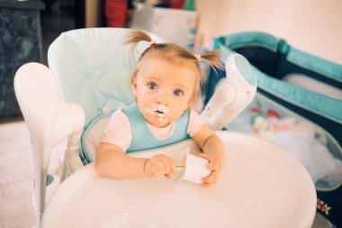 Mutfakta, masada, çocuk sandalyesinde yoğurt yiyen bir kız çocuğu.