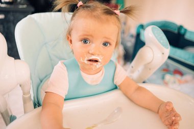 Mutfakta, masada, çocuk sandalyesinde yoğurt yiyen bir kız çocuğu.