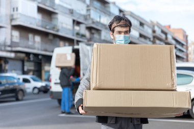 Üniformalı ve maskeli teslimatçı arka planda odak noktasından büyük bir teslimat kutusu ve minibüsü taşıyor.