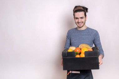 Elinde meyve kutusuyla teslimatçı çocuk siparişi getiriyor, önlük giymiş ve gülümsüyor.