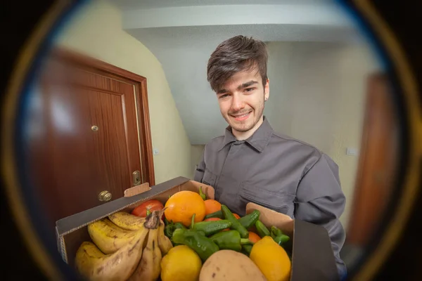 Delivery man with fruit box in his hands delivering the order, dressed in uniform and smiling, customer looking through the peephole