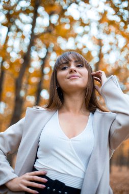 Sonbahar parkında açık renk ceketli, turuncu yapraklı, alttaki manzaralı zarif beyaz kadın.