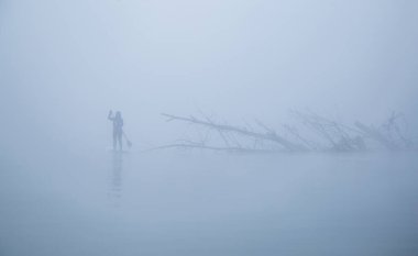 Ağacın yakınındaki yoğun sislerle çevrili nehir kıyısındaki yemek masasında kürek çeken tanınmayan bir kadın.