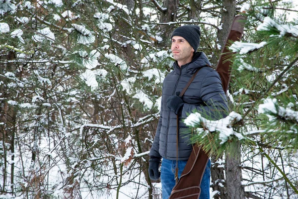 smiling caucasian man in down jacket and jeans with shotgun in the case on the shoulder in the winter forest