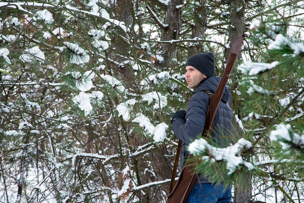 man in down jacket and jeans with shotgun in the case on the shoulder in the snowy forest