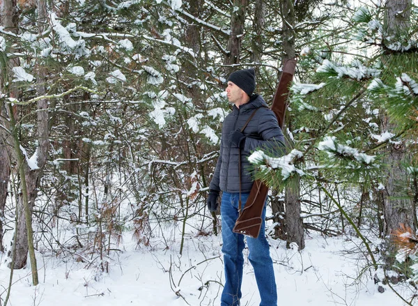 caucasian man in down jacket and jeans with shotgun in the case on the shoulder in the winter forest