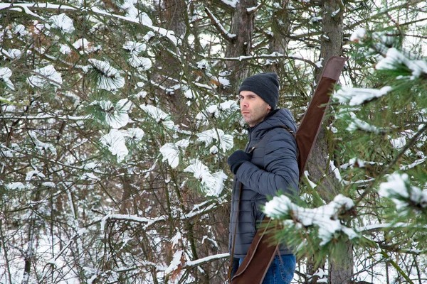 man in down jacket and jeans with shotgun in the case on the shoulder in the snowy forest