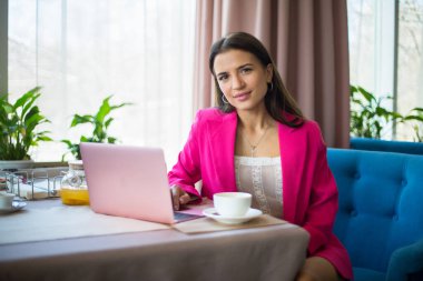 Kırmızı takım elbiseli genç bir kadın restoranda çay ve dizüstü bilgisayarla masada oturuyor.