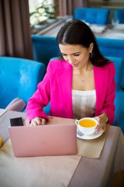 Kırmızı takım elbiseli genç bir kadın restoranda çay ve dizüstü bilgisayarla masada oturuyor.