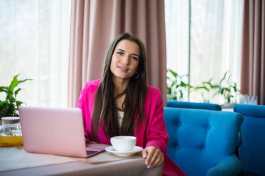 Kırmızı takım elbiseli genç bir kadın restoranda çay ve dizüstü bilgisayarla masada oturuyor.