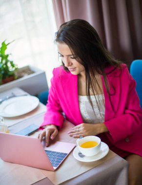 Kırmızı takım elbiseli genç bir kadın restoranda çay ve dizüstü bilgisayarla masada oturuyor.