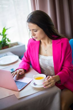 Kırmızı takım elbiseli genç bir kadın restoranda çay ve dizüstü bilgisayarla masada oturuyor.