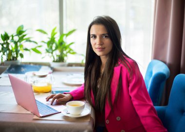 Kırmızı takım elbiseli genç bir kadın restoranda çay ve dizüstü bilgisayarla masada oturuyor.