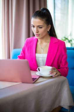 Kırmızı takım elbiseli genç bir kadın restoranda çay ve dizüstü bilgisayarla masada oturuyor.