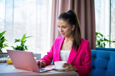 Kırmızı takım elbiseli genç bir kadın restoranda çay ve dizüstü bilgisayarla masada oturuyor.