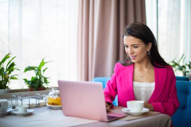 Kırmızı takım elbiseli genç bir kadın restoranda çay ve dizüstü bilgisayarla masada oturuyor.