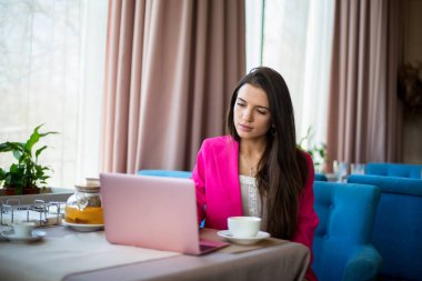 Kırmızı takım elbiseli genç bir kadın restoranda çay ve dizüstü bilgisayarla masada oturuyor.