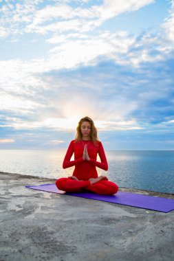 Kırmızı yoga kıyafetli bir kadın nilüferde oturuyor güneş ışınlarında asana 'ya poz veriyor ufukta mavi bulutlar.