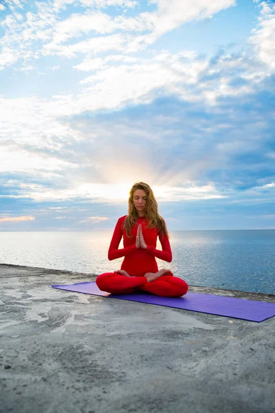 female in a red yoga suit is sitting in lotus pose asana in sun rays near a sea, blue clouds on the horizon