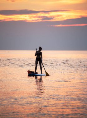 Bir kadın gün doğumunun turuncu ışığında sudaki SUP tahtasının üzerinde kürek çekiyor. Gün doğumuna karşı bir siluet.