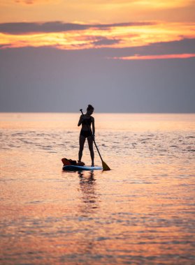Bir kadın gün doğumunun turuncu ışığında sudaki SUP tahtasının üzerinde kürek çekiyor. Gün doğumuna karşı bir siluet.