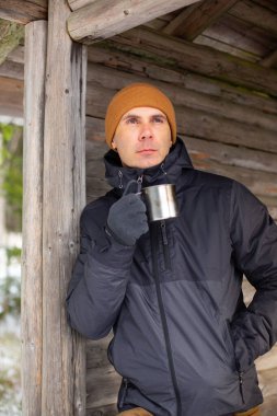 Sıcak bir içecekle ormandaki ahşap bir verandada duran düşünceli beyaz adam.