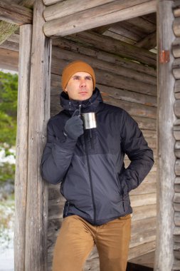 Beyaz ceketli bir adam ahşap bir kulübe duvarına yaslanmış ve metal bir bardaktan su içiyor.