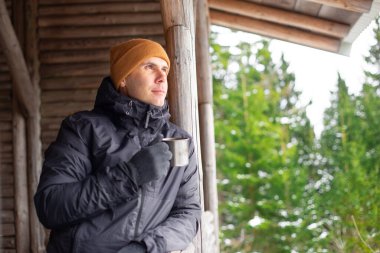 Düşünceli beyaz adam, kalın giysiler içinde, tahta bir orman kulübesinin terasında metal bir bardak tutuyor.