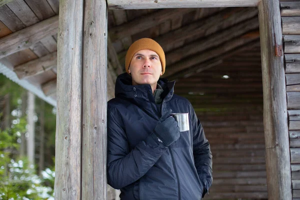 Beyaz ceketli bir adam ahşap bir kulübe duvarına yaslanmış ve metal bir bardaktan su içiyor.