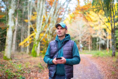 Sonbaharda bir orman yolunda elinde akıllı bir telefonla bekleyen bir adam. Renkli sonbahar yaprakları, ağaçlar ve yumuşak doğal ışık ile açık hava seyrüsefer ve seyahat kavramı