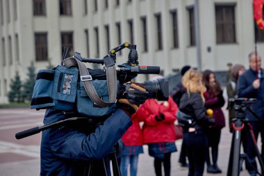 Büyük kameralı bir fotoğrafçı, komünist devrimin yıldönümünde meydanda bayraklarla dikilen insanların videosunu çeker..