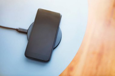 Yuvarlak şarj pedinde duran akıllı telefonun en alt görüntüsü. Açık mavi ve ahşap arkaplan. Modern teknoloji, kablosuz aygıt ve enerji aktarımı.