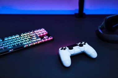 Beyaz oyun kumandası, arka ışıklandırmalı klavye ve gri masadaki kulaklıklar. Profesyonel bilgisayar oyunu, esport işi ve online dünya konsepti.