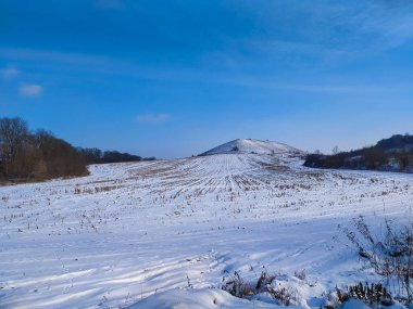 The rural landscape with snow covered field, hill and the blue sky