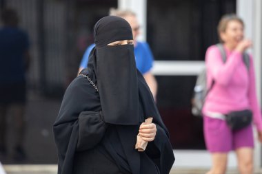 İstanbul, Türkiye - 5 Ekim 2025: Arkaplanda parlak giyimli turistlerle tezat oluşturan ve yüzünü kaplayan siyah yaka takılı bir kadın kamuya açık alanda yürürken görülüyor.