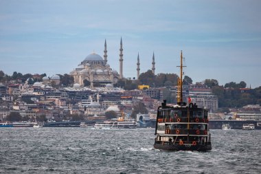 İstanbul, Türkiye - 5 Ekim 2025: Eminonu ilçesi üzerindeki ufuk çizgisinde tarihi Süleyman Camii (Süleyman Camii) egemen olduğu Haliç 'e ait siyah ve sarı geleneksel bir feribot denize açılıyor.