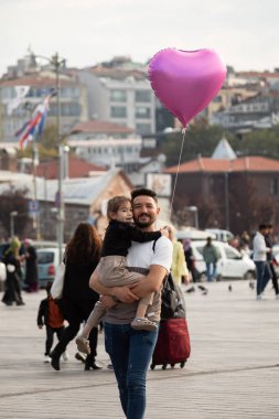 İstanbul, Türkiye - 5 Ekim 2025: Meşgul bir halk meydanında yürürken gülümseyen bir baba, kafalarının üzerinde aşkı simgeleyen, pembe bir kalp şeklinde balon tutuyor..
