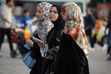 İstanbul, Türkiye - 5 Ekim 2025: Farklı türban (türban) takan ve tek bir kırmızı gül taşıyan üç genç Müslüman kadın, Asya tarafında kalabalık bir halk meydanında yürüyor.