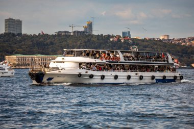 İstanbul, Türkiye - 5 Ekim 2025: Kalabalık bir tur teknesi tarihi Dolmabahce Sarayı 'ndan ve Beşiktaş siluetinin yüksek binalarından geçiyor.